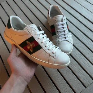 Gucci Ace sneakers med broderad bi - Säljer ett par Gucci Ace sneakers i vitt skinn med klassiska gröna och röda ränder på sidan och en broderad guldfärgad bi. Baksidan har metallicrött och metallicgrönt med Gucci-logga. Platt sula och snörning framtill. Riktigt snygga och ikoniska sneakers för dig som gillar lyx.
