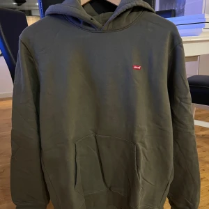 Olivgrön hoodie från Levi's i bomull - Säljer en olivgrön hoodie från Levi's i storlek S. Tröjan har klassisk huva, magficka och liten röd Levi's-logga på bröstet. Tillverkad i mjuk bomull och har långärmad passform med ribbade muddar. Perfekt för chill dagar och streetwear.