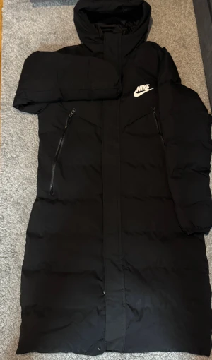 Svart lång pufferjacka från Nike - Svart pufferjacka från Nike med huva och två snedställda dragkedjefickor framtill. Jackan har en clean look med vitt Nike-logga på bröstet och långa ärmar. Perfekt för kalla dagar och har en längre modell som täcker bra. Materialet är polyester.