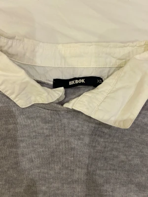 Grå tröja med vit krage från Bik Bok - Supersöt grå tröja från Bik Bok i storlek XS med en vit klassisk skjortkrage. Tröjan har lång ärm och är gjord i mjukt stickat material som känns skönt mot huden. 