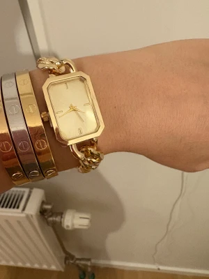 Guldfärgad fyrkantig länk-klocka - Snygg guldfärgad klocka med fyrkantigt ur och chunky länkarmband. Urtavlan är stilren med minimalistiska markeringar och guldfärgade visare. Länkarna är blanka och ger ett lyxigt intryck, perfekt för dig som gillar statement-accessoarer.