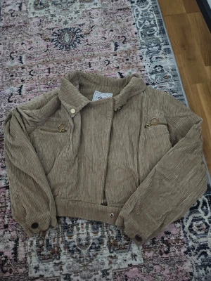 Beige manchester teddyjacka - Säljer en beige teddyjacka i manchester med mjukt teddyfoder. Jackan har guldiga dragkedjor, coolt bälte med metallspänne och ribbade muddar. Croppad passform och långärmad.