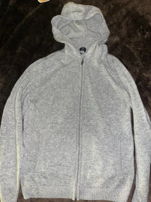 Grå hoodie med dragkedja från Boggi Storlek M - Snygg ljusgrå hoodie från Boggi Milano i cashmere blend. Tröjan har huva, hel dragkedja framtill och två fickor. Ribbstickade muddar vid ärmslut och nederkant ger en schysst passform. Perfekt för chill dagar och när du vill ha det extra mjukt. Storlek M, passar 170-180cm. 