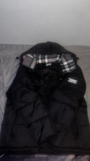 Svart Burberry pufferjacka/väst - Säljer en svart pufferjacka från Burberry med klassisk rutig huva i grått, vitt och rött. Jackan har dragkedja, tryckknappar och Burberry London England-logga på ärmen. Yttermaterialet är syntet och fodret är mjukt och varmt. Perfekt för kalla dagar. Går även ta bort armarna och görs till en väst, Postar inom 24h efter betalning.