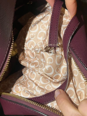 Ryggsäck från Guess - Snygg vinröd ryggsäck från Guess med guldiga dragkedjor och präglad logga framtill. Väskan har ett mönstrat innerfoder med G-loggor och flera fack för smart förvaring. Tillverkad i syntetmaterial med justerbara axelband. Lite slitage, se bilder.