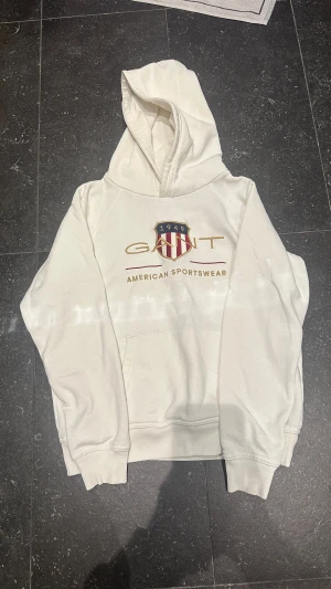 Vit hoodie från GANT med tryck - Vit hoodie från GANT med tryck. Tröjan har huva, magficka och ribbade muddar. Materialet är mjuk bomull och passformen är normal. Perfekt för chill dagar och sportig stil. Tröjan är varsamt använd och det finns inga defekter, ser ut som ny! Storlek 158-164cm.☺️
