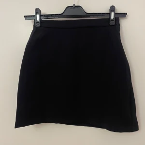 Svart kjol i kostym material  - En stilren svart kostym kjol med rak passform och enkel design. Kjolen har en bred linning och är tillverkad i ett mjukt, tjockare bomullstyg som känns skönt mot huden. Perfekt för dig som gillar minimalistiska plagg och vill ha en klassisk look. Är ganska stor i storlek så passar mer en 36 skulle jag säga.