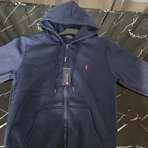 Marinblå hoodie från Polo Ralph Lauren - Snygg marinblå hoodie från Polo Ralph Lauren med röd broderad logga på bröstet. Tröjan har huva med snörning, dragkedja framtill och två praktiska fickor. Mjuk insida i fleece och klassisk passform, perfekt för chill dagar.