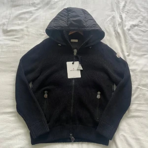 Svart pufferjacka från Moncler - Svart pufferjacka från Moncler med stickade ärmar och ribbade muddar. Jackan har quiltad huva och rygg, två dragkedjeförsedda fickor fram och Moncler-logga på ärmen. Materialmix av polyester och stickad ull ger en schysst look och grym komfort.