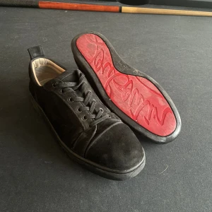 Svarta sneakers från Christian Louboutin - Säljer ett par svarta sneakers från Christian Louboutin med klassisk röd sula och signaturtext. Skorna har rund tå, snörning och är tillverkade i mocka med svart yttermaterial och beige insida. Perfekt för dig som vill sticka ut med exklusiva detaljer. Är använda men inte något som är trasig eller inte går att tvätta bort.