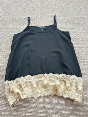 Svart linnetopp med spets från Romeo & Juliet - Svart linnetopp från Romeo & Juliet Couture i storlek S. Toppen har tunna axelband och en bred, beige spetskant med blommigt broderi längst ner. Materialet är mjukt och lätt, perfekt för att skapa en chic look. Spetskanten ger en extra detalj som sticker ut.