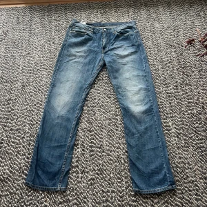 Levi's 514 blå jeans W35 L34 - Säljer ett par klassiska Levi's 517 jeans i blå tvätt med raka ben och bootcut-snitt. Jeansen har fem fickor, knappgylf och snygga slitningar på låren. Perfekt för dig som gillar tidlös denimstil och vill ha ett par jeans med lite extra attityd.