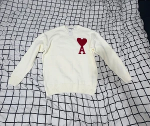 Vit stickad tröja med rött hjärta - Vit stickad tröja från AMI med ett rött hjärta och bokstaven A broderat på bröstet. Tröjan har rund hals, ribbade muddar och långärmad passform. Perfekt för dig som gillar stilrena detaljer med en twist.