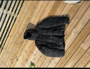 Svart pufferjacka med huva från Hugo boss - Svart pufferjacka från Hugo boss med stor huva och snörning i nederkant. Jackan har quiltad design, dragkedja och knappar framtill samt två sidofickor. Perfekt för kalla dagar och har en loose passform som ger en chill vibe.