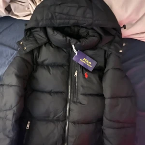 Svart pufferjacka från Polo Ralph Lauren - Svart pufferjacka från Polo Ralph Lauren med huva och två sidofickor med dragkedja. Jackan har en röd Polo-logga broderad på bröstet och snygga quiltade paneler. Materialet är syntet och jackan stängs med dragkedja framtill.