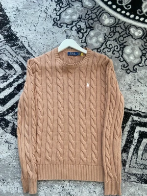 Beige kabelstickad tröja Polo Ralph Lauren - Snygg beige kabelstickad tröja från Polo Ralph Lauren med klassisk rund hals och diskret broderad logga på bröstet. Tröjan har långa ärmar och ribbade muddar, perfekt för dig som gillar stilren och tidlös design.