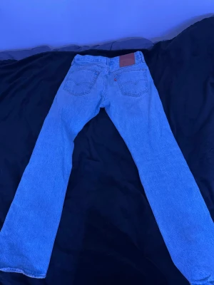 Levi's 501 ljusblå jeans straight fit - Klassiska Levi's 501 jeans i ljusblå tvätt med rak passform. Byxorna har fem fickor, knappgylf och det ikoniska läderpatchen bak i midjan. Perfekta för dig som gillar tidlös stil och vill ha ett par jeans som funkar till allt.