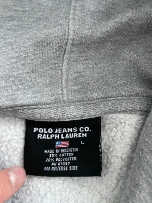 Grå hoodieRalph Lauren - Säljer en grå hoodie från Polo Jeans Co. Ralph Lauren i storlek L. Tröjan har stor vit broderad logga på bröstet, snörning vid huvan och är tillverkad i mjuk bomull och polyester. Perfekt för chill dagar och har klassisk loose passform.