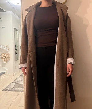 Brun lång kappa med bälte - Säljer en snygg brun kappa i lång modell med klassisk krage och bälte i midjan. Kappan har en stilren look och är tillverkad i ett mjukt ullmaterial. Perfekt för dig som vill ha en chill och tidlös vibe i höst och vinter.
