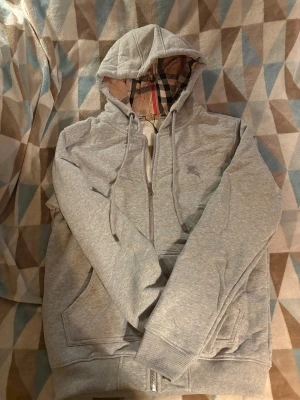 Grå Burberry hoodie med dragkedja - Säljer en grå hoodie från Burberry i storlek M. Tröjan har klassisk Burberry-rutig huva, broderad logga på bröstet, snörning och dragkedja framtill. Mjuk bomullskänsla och ribbade muddar vid ärmar och nederkant. Perfekt för chill dagar. passar S-M