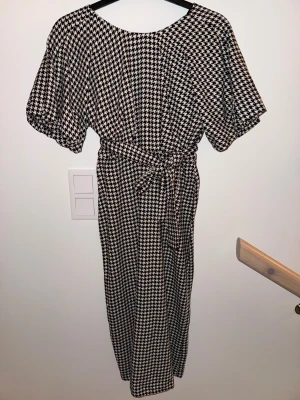 Långklänning med hundtandsmönster och knyt från Zara - Snygg långklänning med svartvitt hundtandsmönster och knytband i midjan. Klänningen har korta ärmar och en avslappnad passform som ger en cool och trendig look. Perfekt för dig som gillar grafiska mönster och vill sticka ut.