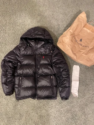 Polo Ralph Lauren glossy jacka (kvitto finns) - Säljer denna riktigt feta polo Ralph lauren jackan i svart färg. Den är köpt från Göteborg som ni ser så förekommer kvitto samt orginal påsen. Den är storlek S och Perfekt för kalla dagar och har en clean, modern look. Den är i bra skicka finns ett väldigt litet hål på jackan.
