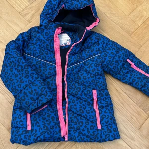 Blå leopardmönstrad pufferjacka - Blå pufferjacka med svart leopardmönster och rosa detaljer på dragkedjor och huva. Jackan har huva, långa ärmar och flera fickor med dragkedja. Perfekt för kalla dagar och sticker ut med sitt djurmönster.