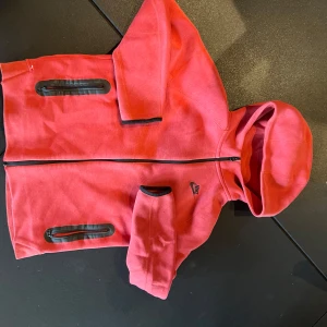 Röd hoodie från Nike med dragkedja XL - Nike hoodie i rött med svart logga på bröstet. Tröjan har hel dragkedja, huva och svarta detaljer vid fickorna. Tillverkad i mjukt bomullsmaterial och har långa ärmar. Perfekt för chill eller träning.