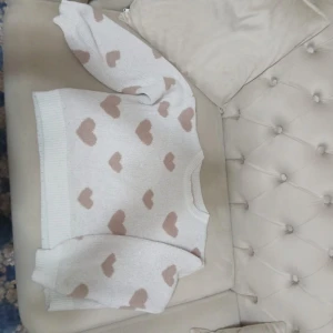 Vit stickad tröja med hjärtan - Supergullig vit stickad tröja med beiga hjärtan över hela. Tröjan har rund halsringning, ribbade muddar och är långärmad. Perfekt för dig som vill ha en mysig och söt look med lite extra kärlek i outfiten. Skulle någon säga för barn men funkar för damer också:)