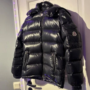 Moncler jacka - Säljer en svart, glansig pufferjacka från Moncler med huva och dragkedja. Jackan har Moncler-logga på ärmen, två fickor med dragkedja och en schysst, vadderad look. Perfekt för kalla dagar när du vill hålla stilen och värmen.