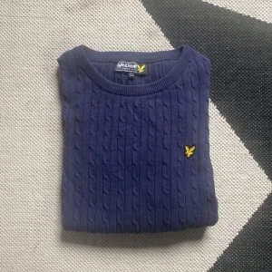 Marinblå kabelstickad Lyle & Scott tröja - Kabelstickad marinblå tröja från Lyle & Scott med rund hals och gul logga broderad på bröstet. Tröjan har långa ärmar och ribbade muddar. Perfekt för lager-på-lager under hösten och vintern.  Har väldigt litet slitage på vänstra armkragen som bara ses på närhåll.                                                                  Storleken är runt 164-170cm junior.