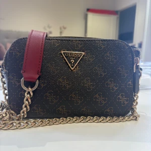 Guess brun axelväska med kedja - Snygg brun axelväska från Guess med guldiga detaljer och kedjerem. Väskan har Guess-logga framtill, mönstrad yta och två dragkedjefack. Axelremmen har en vinröd läderdetalj för extra stilpoäng. Perfekt storlek för det viktigaste. Köptes för 800