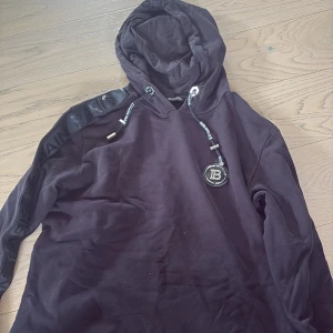 Svart hoodie från Balmain Paris - Svart hoodie från Balmain Paris med huva, långa ärmar och coola dragkedjor i sidorna. Axelband med Balmain Paris-tryck och snyggt B-emblem framtill. Mjuk bomullskvalitet och detaljer i silver. Perfekt för dig som vill sticka ut.