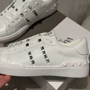 Valentino Rockstud vita sneakers - Snygga vita Valentino Garavani Rockstud sneakers med silverfärgade nitar och klassisk snörning. Skorna är i skinn och har en chunky vit sula med mönstrad undersida. Perfekt för dig som vill sticka ut med exklusiva detaljer och stilren design.