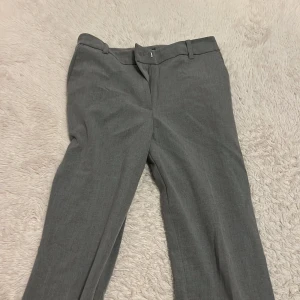 Grå kostymbyxor från BikBok, stl 36 - Snygga grå kostymbyxor från BikBok i klassisk rak modell med hällor för bälte och pressveck. Byxorna har en stilren look och passar perfekt till en dressad outfit. Materialet känns mjukt och följsamt, vilket gör dem bekväma hela dagen. Använda 1 gång och kommer ej till användning mer