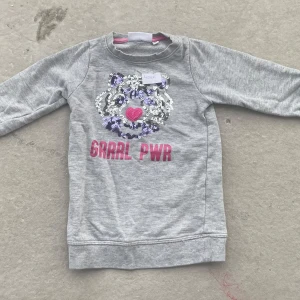 Grå tröja Sweet Heart med paljetter - Grå långärmad tröja från Sweet Heart med paljettdekorerat tigeransikte i silver, lila och rosa samt texten 'GRRRL PWR' i rosa framtill. Mjuk mudd i hals, ärmslut och nederkant. Perfekt för dig som gillar statement-plagg!