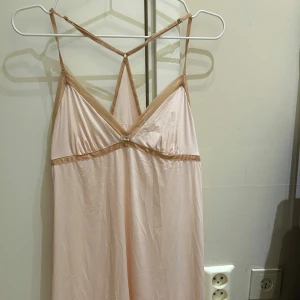 Ny Ljusrosa Triumph nattlinne med smala axelband - Supersöt ljusrosa nattlinne  från Triumph med smala axelband som korsas i ryggen. Nattlinne  har v-ringning, glansiga kantband och en liten rosett framtill. 