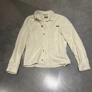 Vit manchesterjacka från Nudie Jeans - Snygg vit manchesterjacka från Nudie Jeans med klassisk skjortkrage, två bröstfickor en på insidan och en på utsidan och knappar framtill. Jackan har lång ärm och en clean, stilren look som passar perfekt till jeans eller chinos.