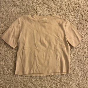 Beige ribbad t-shirt från H&M - En stilren beige t-shirt från H&M med ribbad struktur och klassisk rund hals. T-shirten är kortärmad och har en enkel, clean look som passar till det mesta. Perfekt för dig som gillar en basic vibe och vill ha något som funkar året runt. Ganska liten i storleken