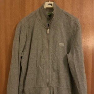 Grå ziptröja från Hugo Boss - Snygg grå ziptröja från Hugo Boss med diskret logga på bröstet. Tröjan har hög krage, hel dragkedja och långa ärmar. Perfekt för en clean och stilren look.