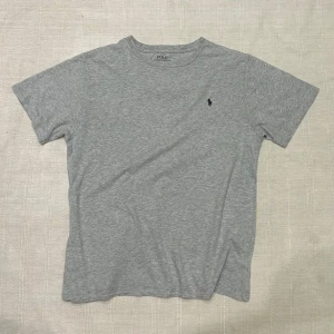 Grå t-shirt från Polo Ralph Lauren - Klassisk grå t-shirt från Polo Ralph Lauren i mjuk bomull. T-shirten har rund hals, korta ärmar och den ikoniska broderade loggan på bröstet. Perfekt basic-plagg med stilren och enkel design.