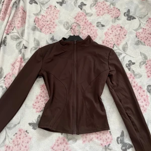 Brun träningsjacka  - Snygg brun träningsjacka med figurnära passform och hel dragkedja framtill. Jackan har hög krage och långa ärmar, perfekt för dig som gillar en stilren och sportig look. Materialet känns mjukt och stretchigt, vilket gör den bekväm att bära.