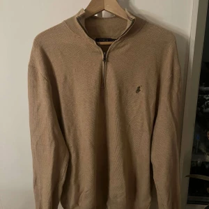 Beige stickad tröja från Polo Ralph Lauren - Säljer en beige stickad tröja från Polo Ralph Lauren med half zip och klassisk broderad logga på bröstet. Tröjan har lång ärm och ribbade muddar vid ärmslut och nederkant. Perfekt för lager-på-lager under hösten och vintern.