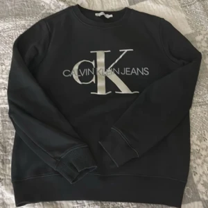 Svart sweatshirt från Calvin Klein Jeans - Svart sweatshirt från Calvin Klein Jeans med stor vit CK-logga framtill. Klassisk rund halsringning och långa ärmar med ribbade muddar. Perfekt för en chill och stilren look. Tillverkad i mjuk bomullsblandning för extra komfort.