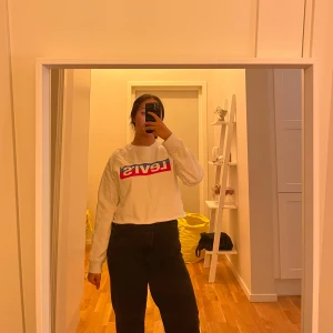Vit croppad Levi's sweatshirt - Säljer en vit croppad sweatshirt från Levi's med färgglad logga framtill. Tröjan har lång ärm och en relaxed passform, perfekt för en chill och trendig look. Materialet känns mjukt och skönt mot huden.