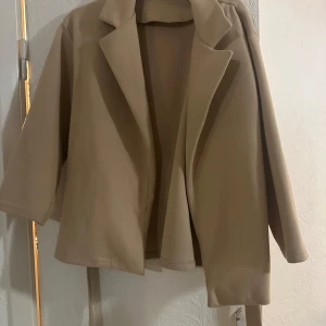 Beige kappa från Missay med bälte - Stilren beige kappa från Missay med klassisk krage och bälte i midjan. Jackan har en rak passform och är tillverkad i ett mjukt material som känns lyxigt. Perfekt för dig som gillar en clean och enkel look.