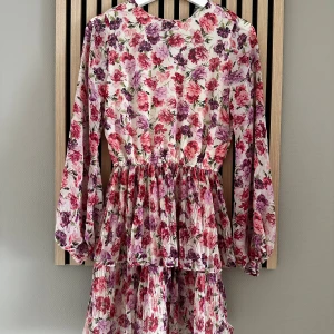 Blommig volangklänning med långa ärmar - Supersöt kort blommig klänning med volanger och långa ärmar. Klänningen har ett mönster i rosa, lila och röda toner på vit botten, markerad midja och lätt rynkade detaljer. Superfin 💕 Mycket finare på än på bild! Endast testad, tyvärr lappen avdragen. 
