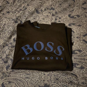 Svart tröja från Hugo Boss med blå logga - Svart långärmad tröja från Hugo Boss med stor blå BOSS-logga framtill. Klassisk rund halsringning med blå detalj på insidan av kragen. Perfekt för dig som gillar stilrena och snygga märkesplagg.