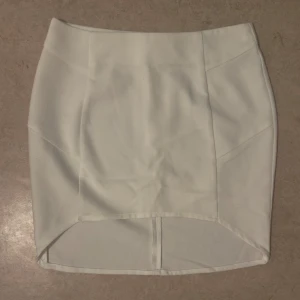 Vit kortkjol med dragkedja - Snygg vit kortkjol med rak passform och markerade sömmar. Kjolen har en silvrig dragkedja bak och är tillverkad i ett slätt tyg som ger en stilren look. Perfekt för dig som gillar minimalistisk stil och vill ha en kjol som sticker ut med sin enkla design.