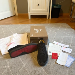Svarta  Christian Louboutin sneakers med nitar - Säljer ett par svarta sneakers från Christian Louboutin med ikonisk röd sula och svarta nitar på tån. Skorna har snörning, är tillverkade i mocka och har en låg siluett. Insidan är fodrad i beige och de levereras med originalkartong och dustbag. Kvitton och tillbehör medföljer. 3 spikar har tyvär försvunnit, se på bild.